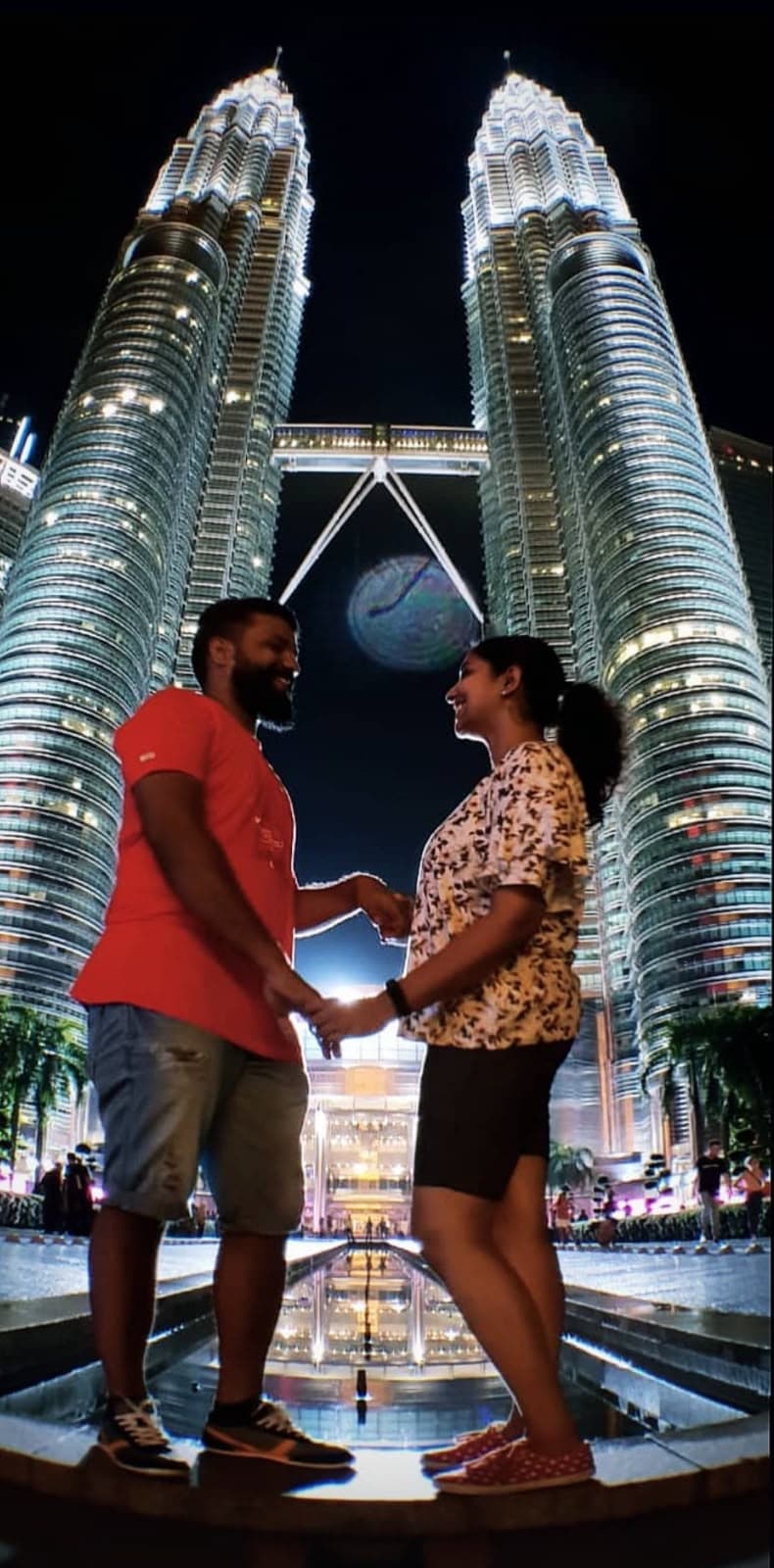 A couple holding hands and smiling at each other in front of the Petronas Towers in Kuala Lumpur, Malaysia, at night